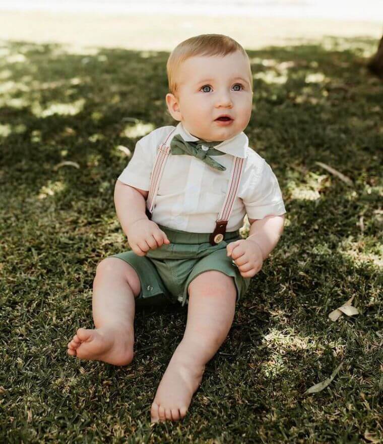 Baby boy sitting on grass, wearing our gentleman suspender shorts and romper outfit in green
