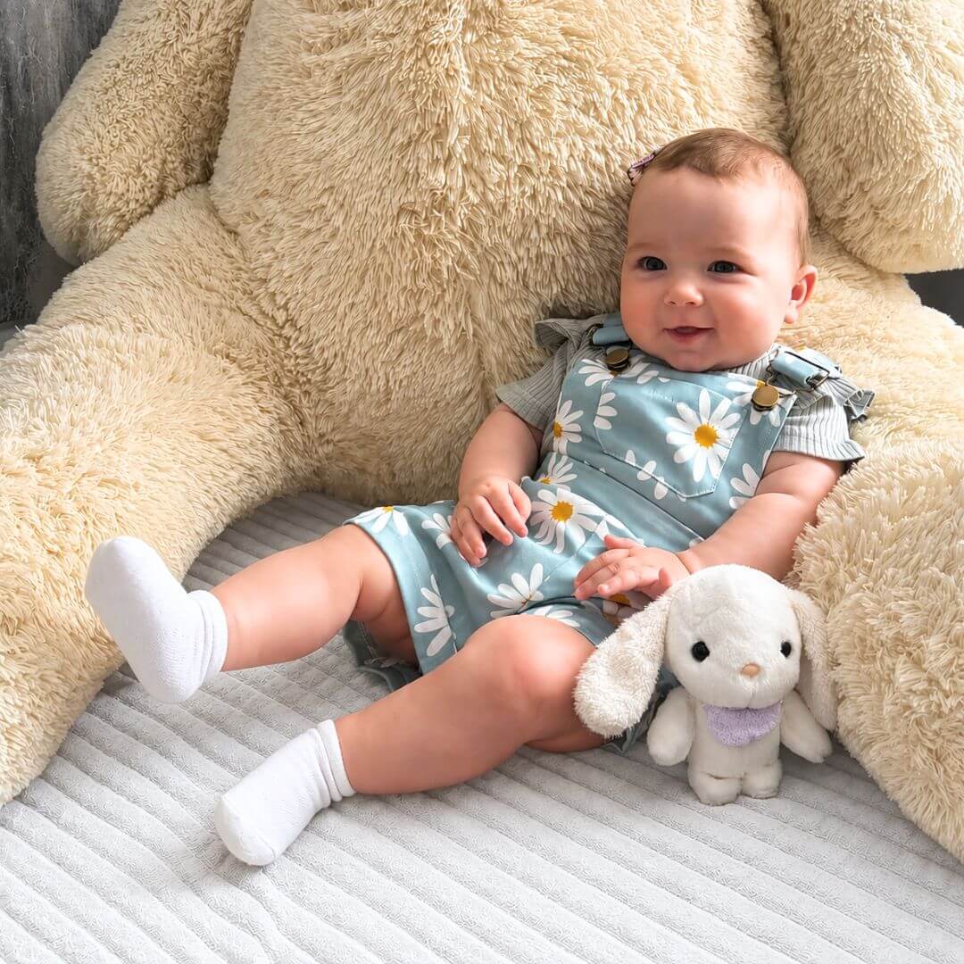 Baby girl wearing blue daisy overall set, sitting on a bed with giant teddy