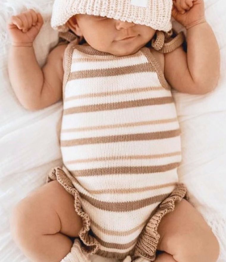 Baby girl in a striped knitted romper with adjustable tie shoulders and ruffle details, wearing a matching hat.