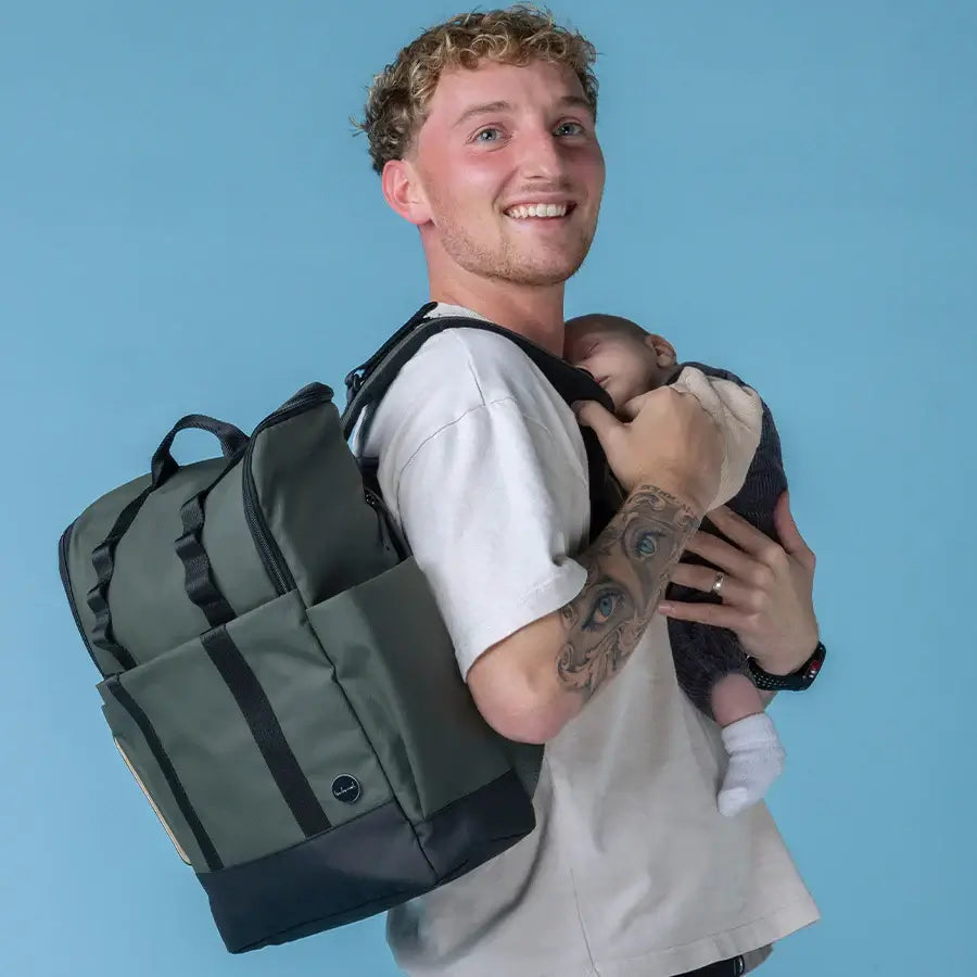 A dad carrying Babymel Finn nappy backpack in olive while holding a baby, showcasing the unisex nappy bag design.