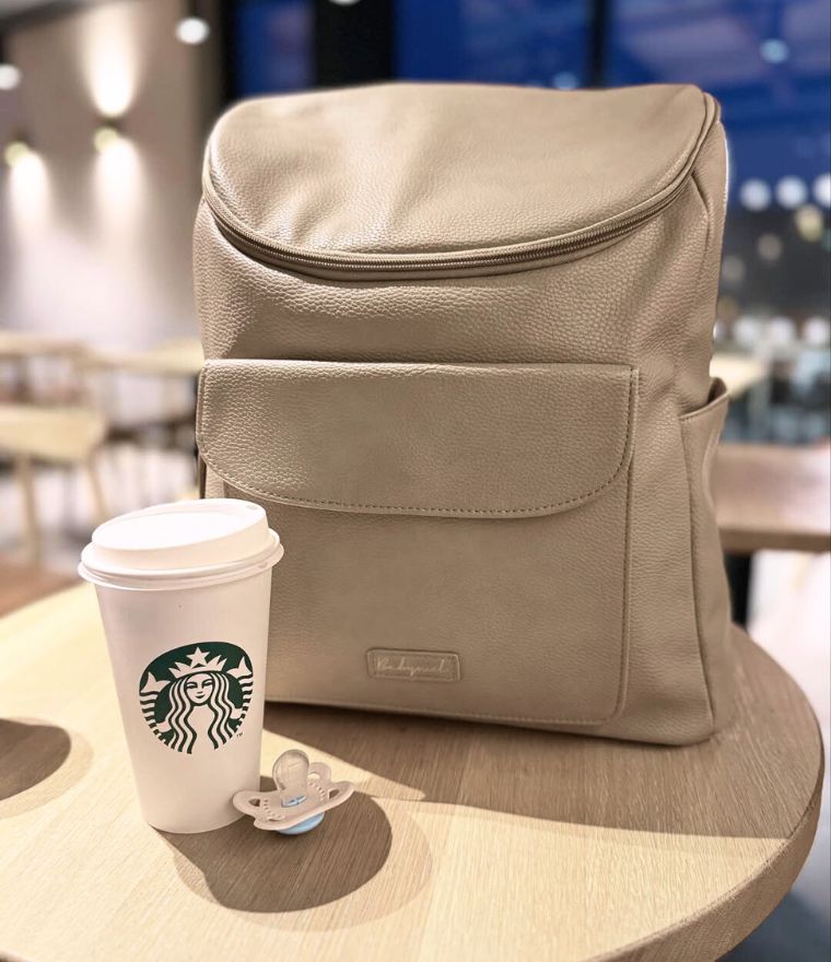 Babymel Lennox nappy backpack in oat, pictured next to Starbucks cup in a cafe