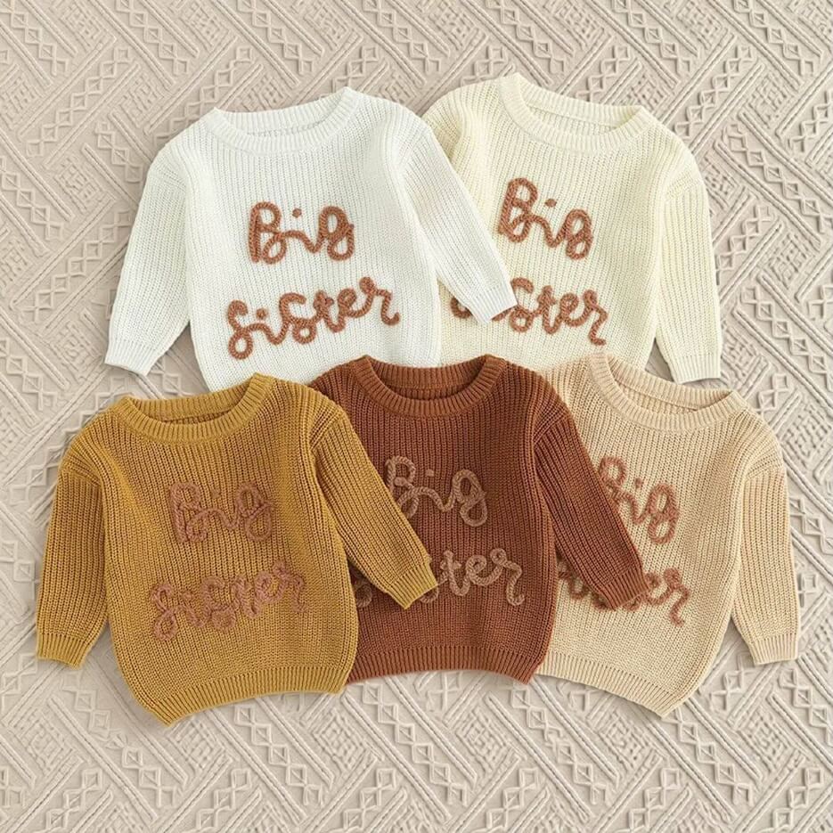 Five big sister knit jumpers in a range of colours, arranged on a textured blanket.