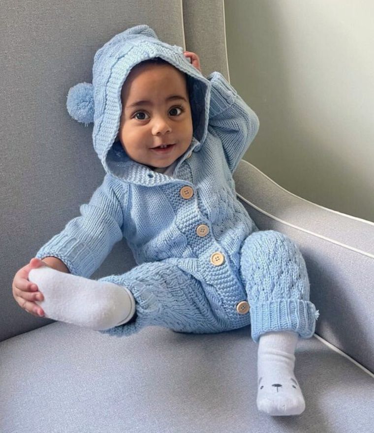Candid shot of a baby boy sitting on a chair wearing our knit baby bear onesie in blue