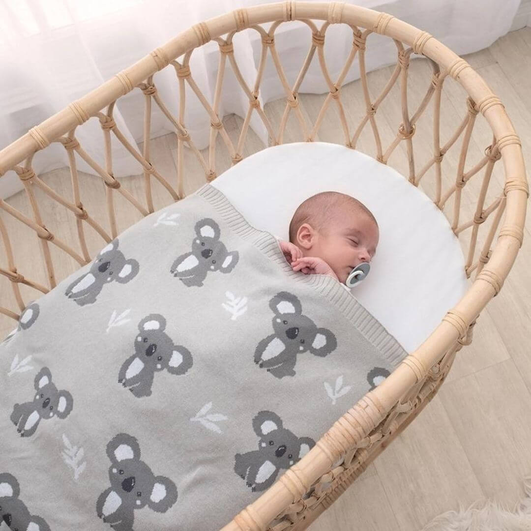 Baby peacefully sleeping in a bassinet, wrapped in a grey koala baby blanket made of 100% cotton knit.