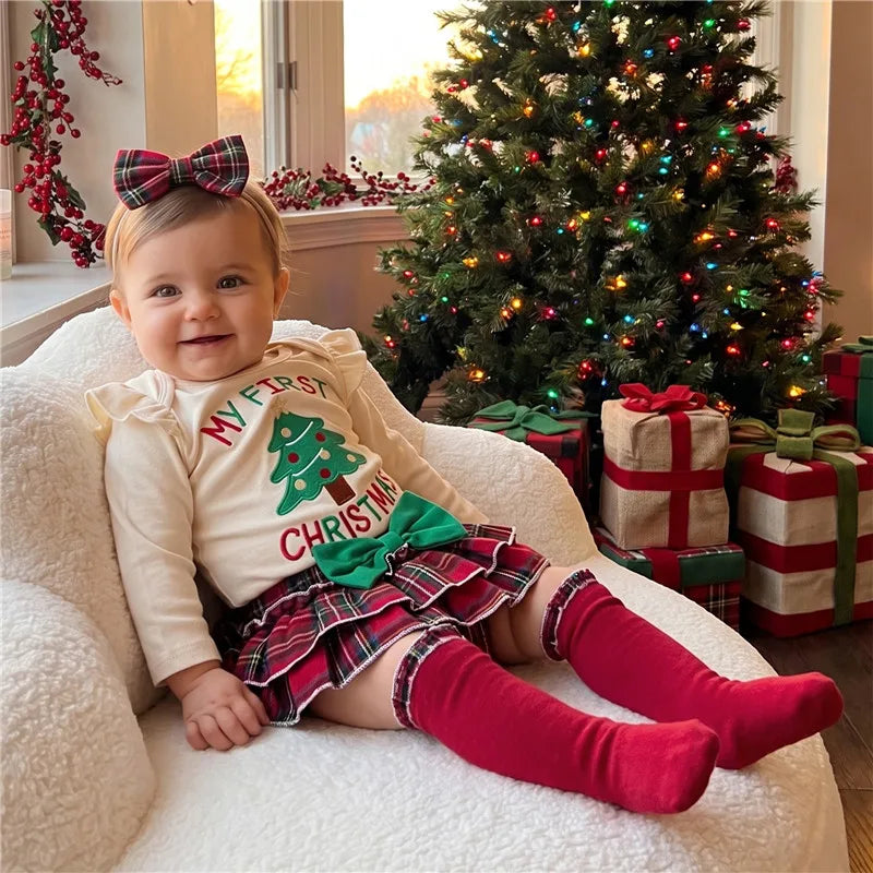 Baby girl sitting on a white armchair near Christmas tree, wearing a 'My First Christmas' baby outfit from Lulu Babe with headband and knee high socks