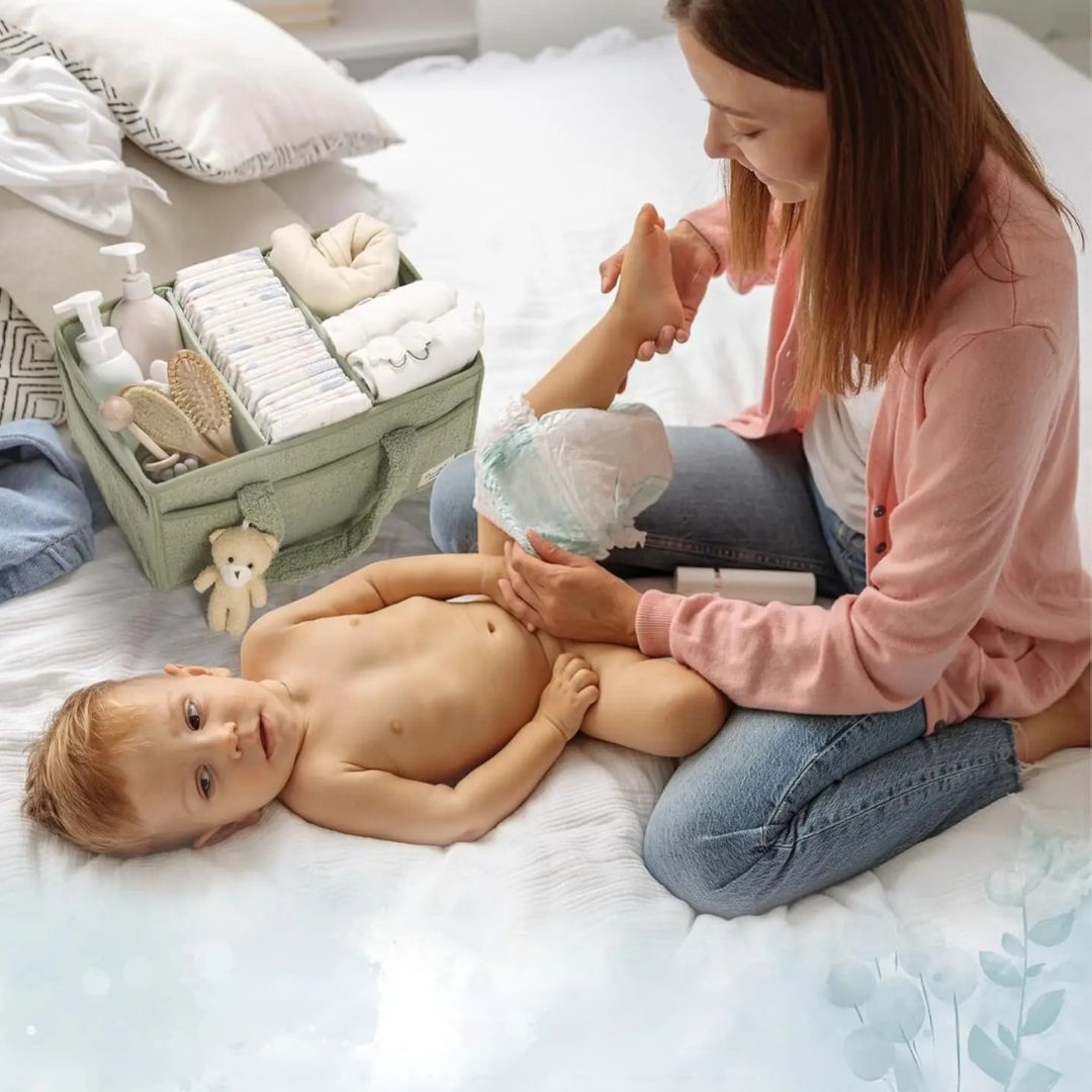 A mother changing a baby's nappy using a green Plush Nappy Caddy for organised baby essentials on a bed.