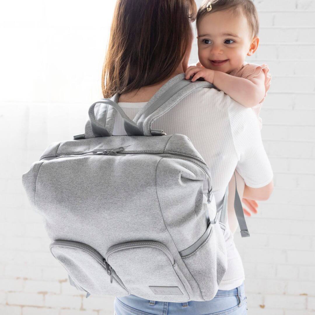 A mum wearing grey neoprene nappy backpack, holding a baby