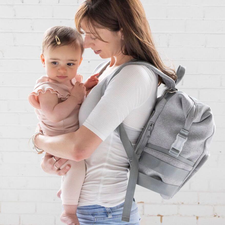 A mother wearing her grey neoprene nappy backpack, holding a baby - side view