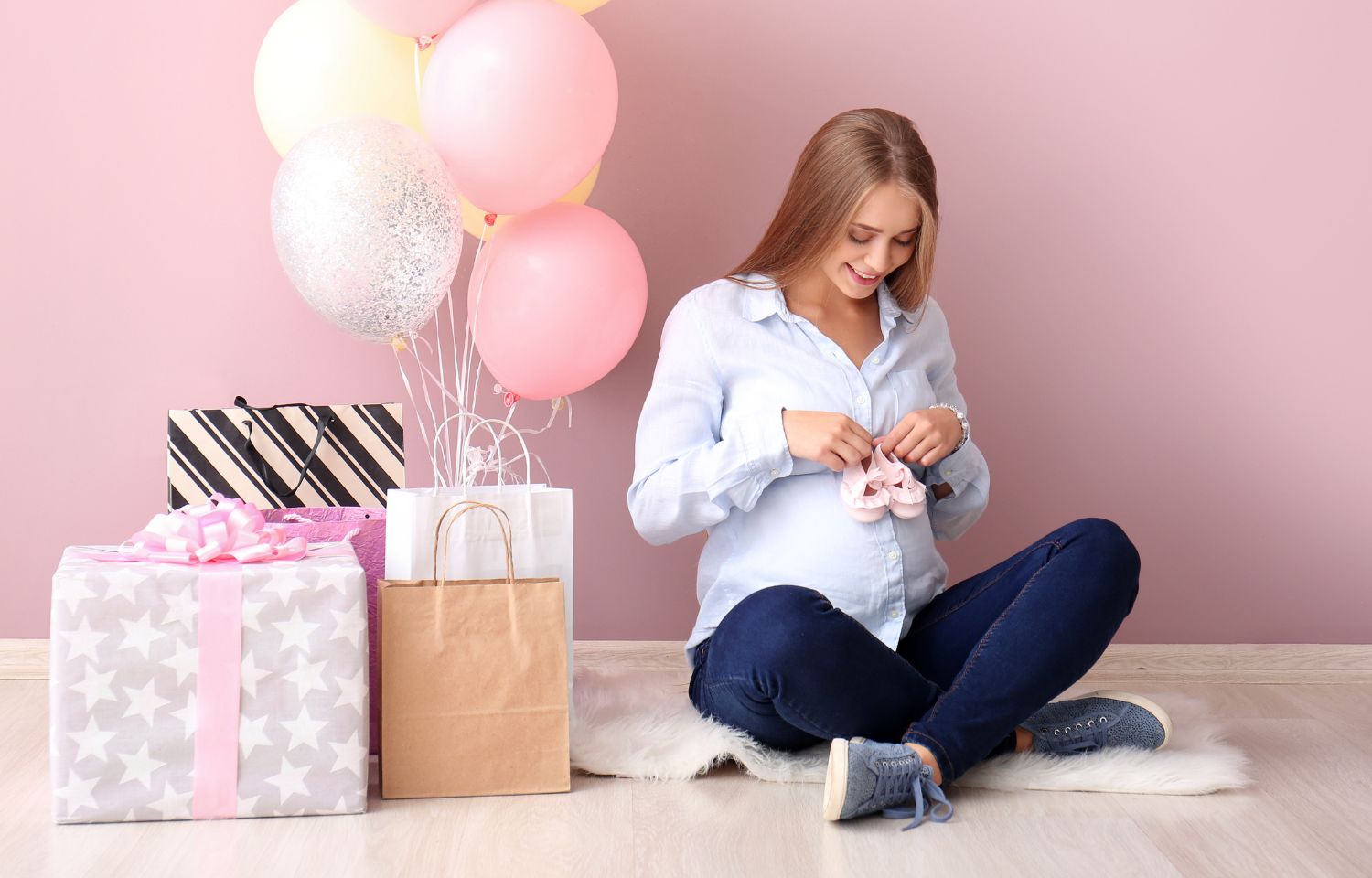 Pregnant woman holding baby shoes, sitting on floor surrounded by baby shower gifts and balloons, celebrating the excitement of expecting a baby.