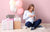 Pregnant woman holding baby shoes, sitting on floor surrounded by baby shower gifts and balloons, celebrating the excitement of expecting a baby.