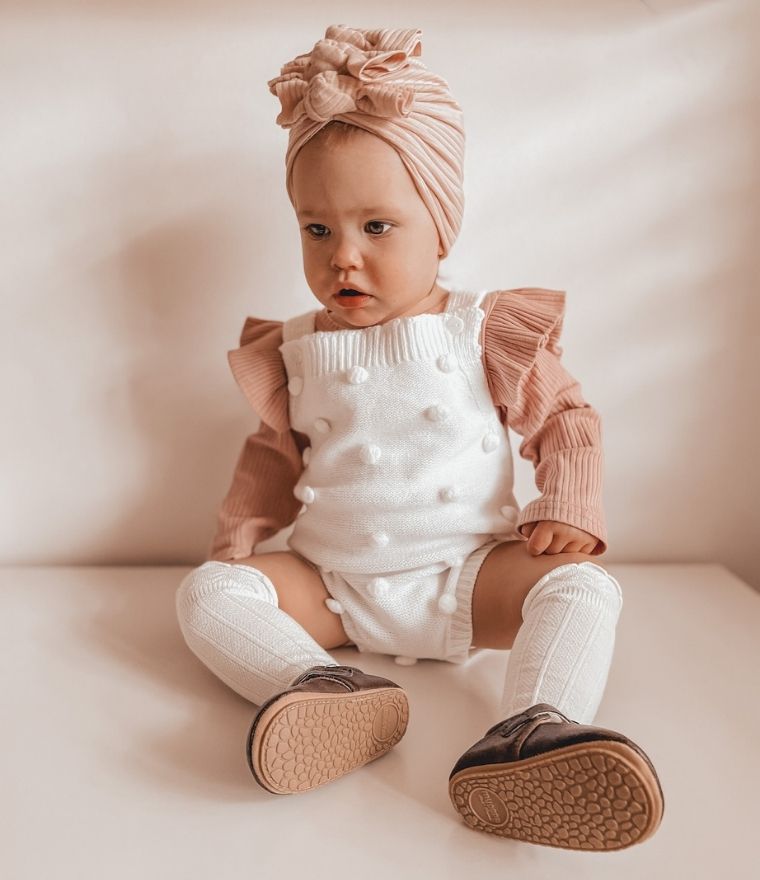 Adorable baby girl wearing a stylish Lulu Babe outfit with a pink turban headband, white ruffled top, and brown shoes, showcasing cute children's clothing.