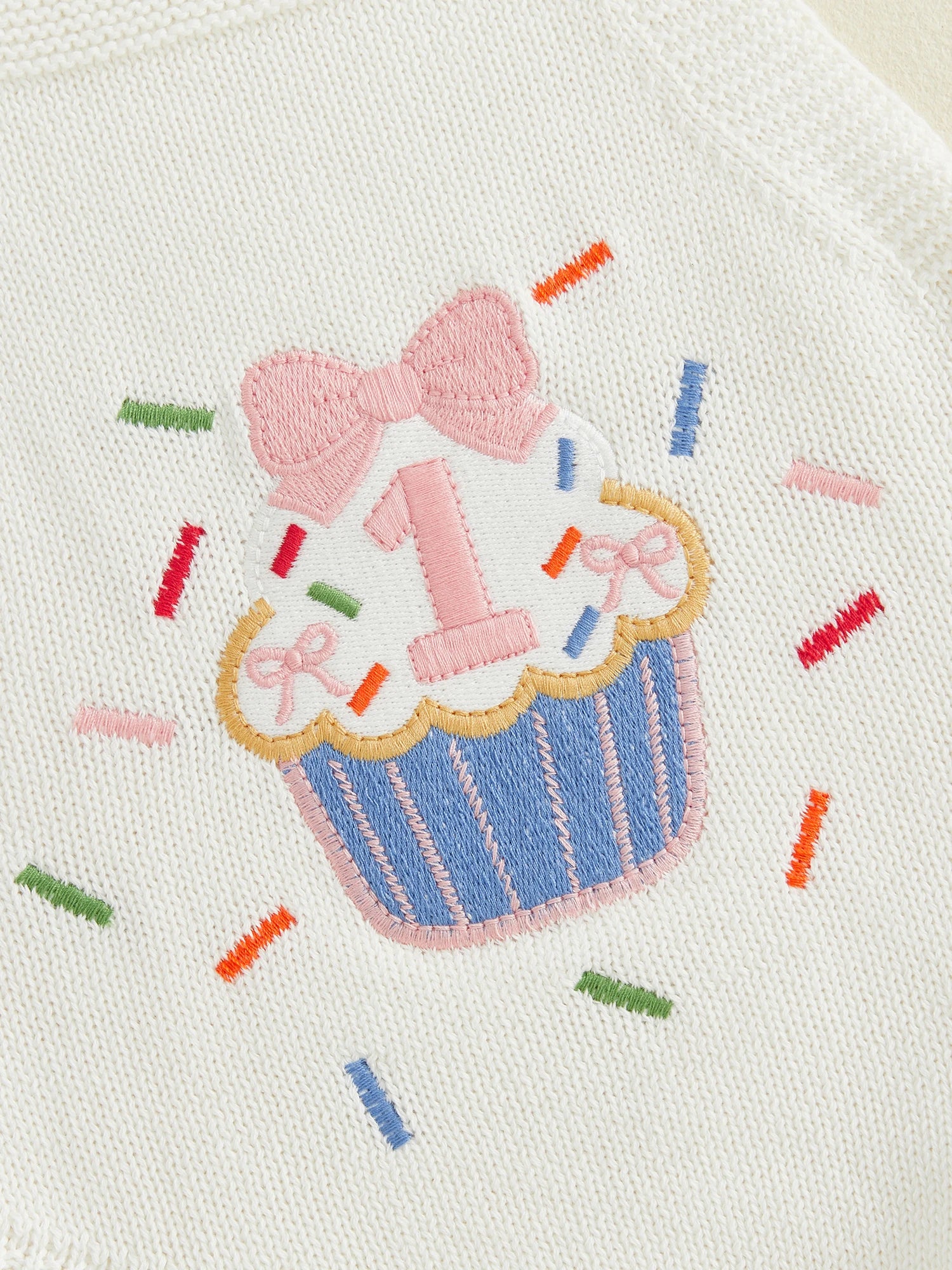 Close-up of embroidered cupcake with "1" detail, bow and rainbow sprinkle stitching on white knit fabric