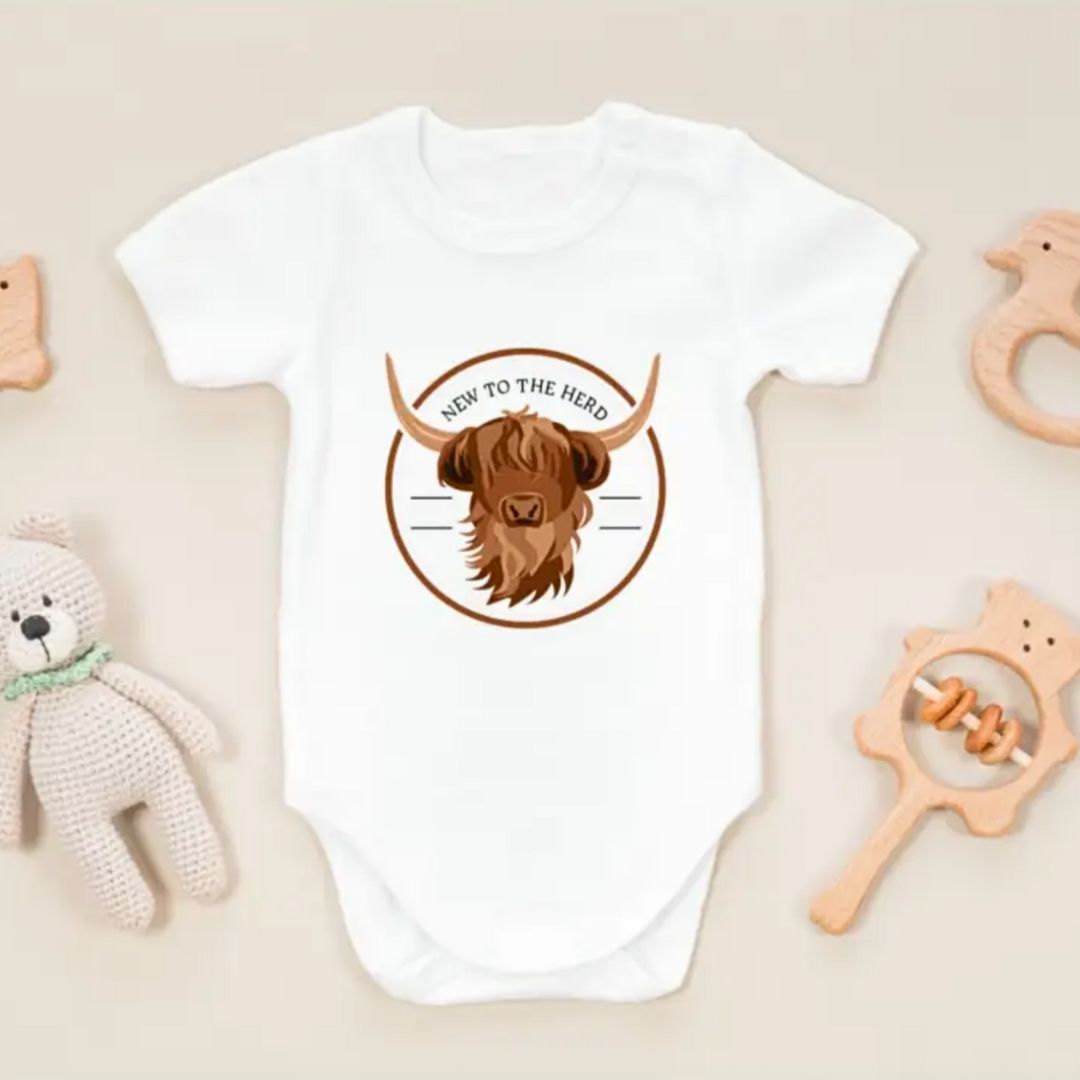 White baby onesie with highland cow graphic and "New to the Herd" text styled flat lay