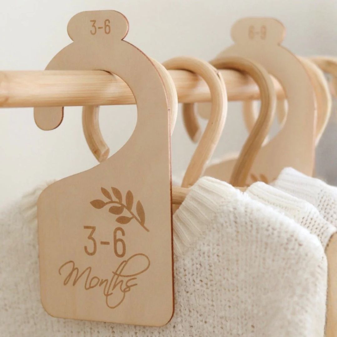 Wooden baby wardrobe divider reading "3–6 Months" with botanical fern engraving hanging on nursery clothing rail between white knit garments