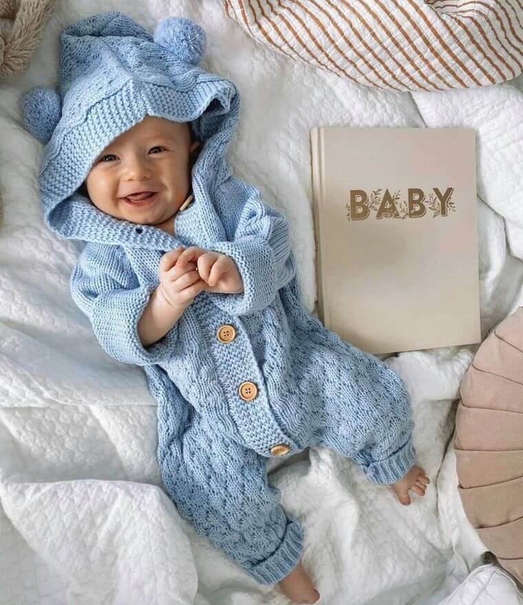 Happy baby wearing a blue cable knit romper with bear ear hood lying on bed