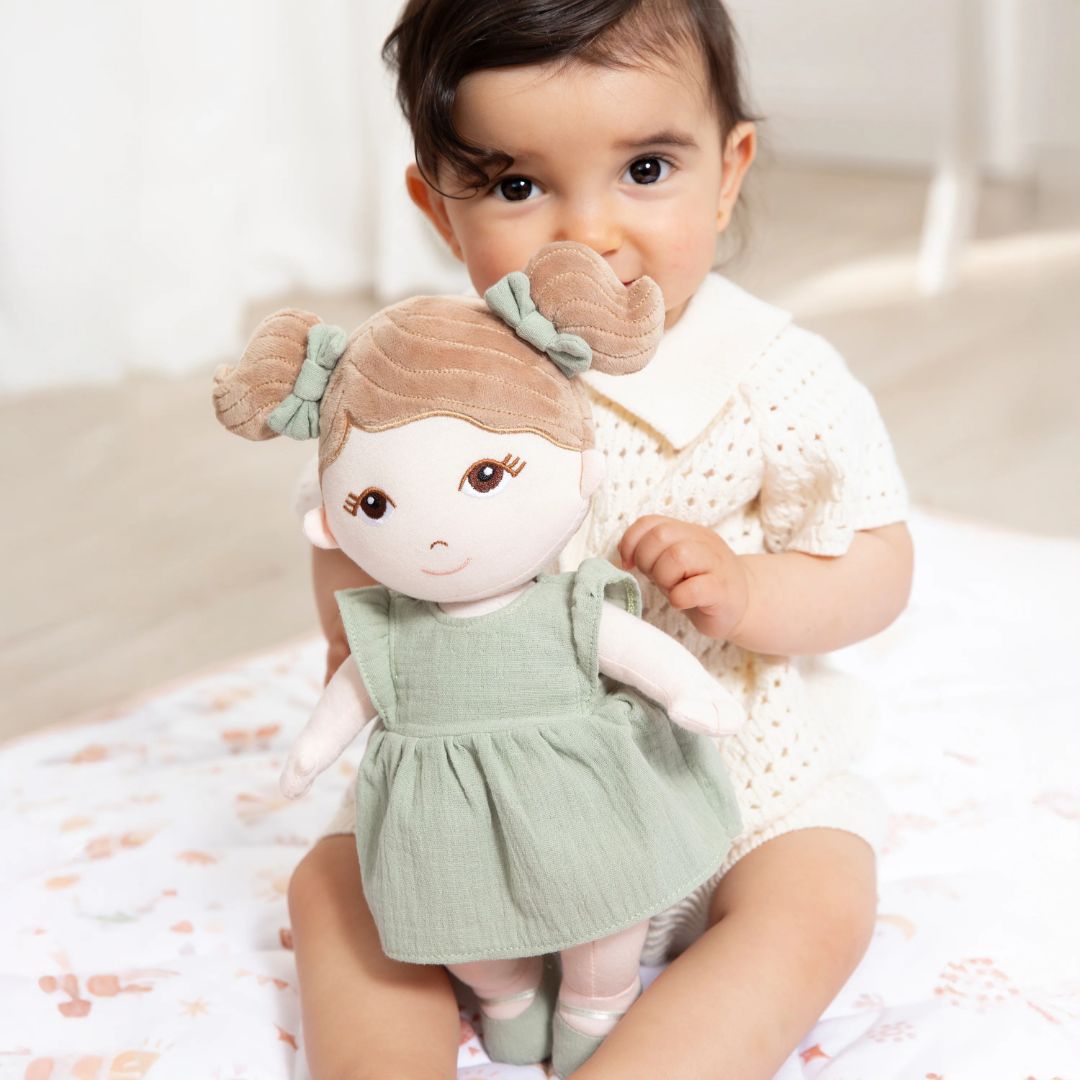Cute toddler holding her soft doll with brown pigtails and a sage green dress