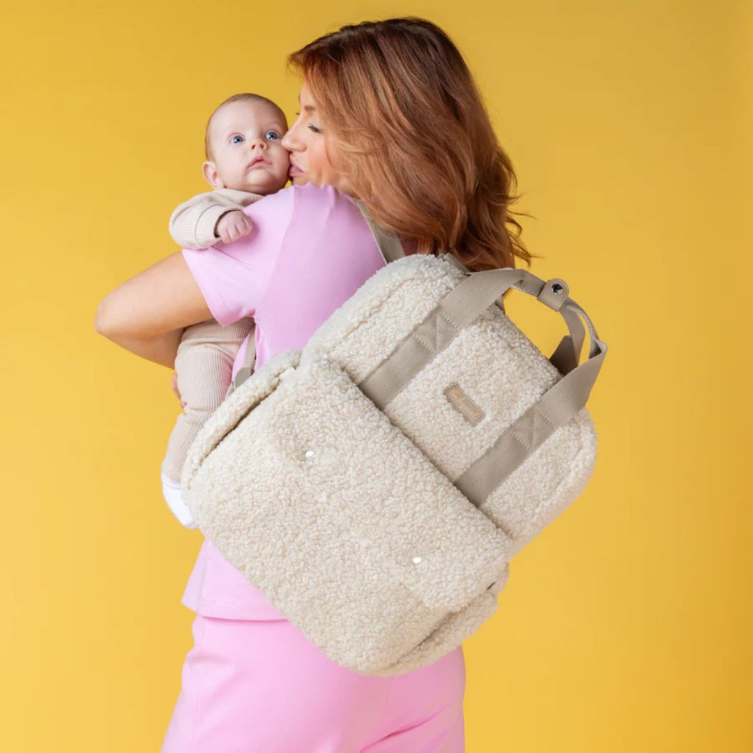 Mum carrying Babymel Teddy Nappy Backpack while holding a baby, showcasing stylish and functional design.