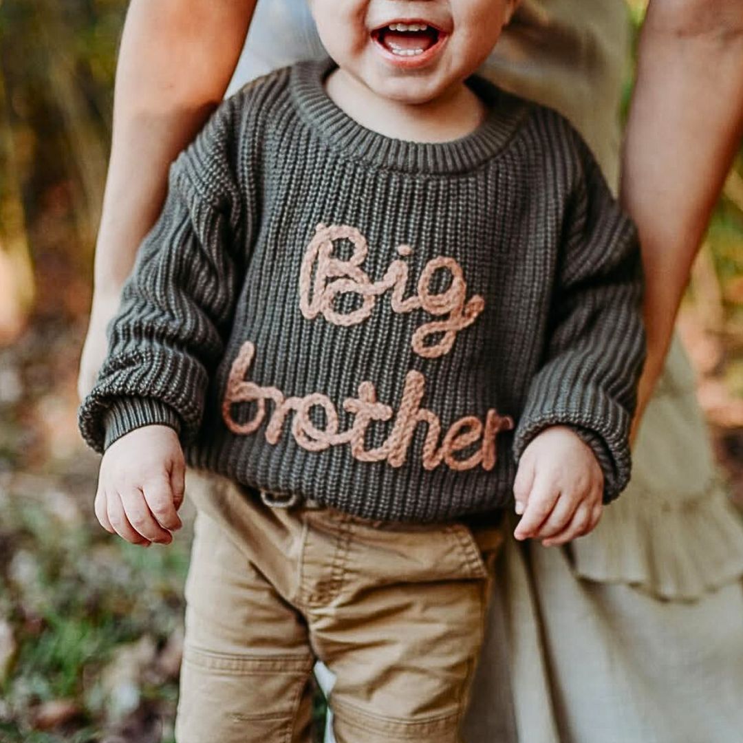Toddler boy wearing a big brother jumper in dark green, standing outdoors.