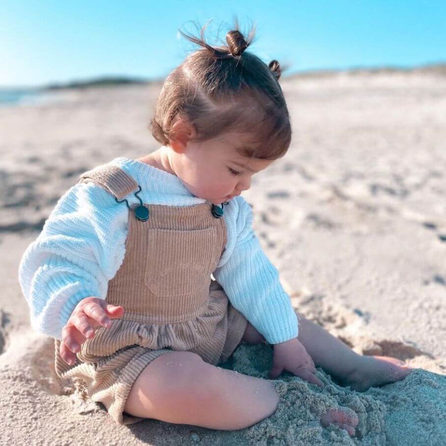 Toddler girl on a sandy beach wearing an oversized white knitted jumper with corduroy overalls