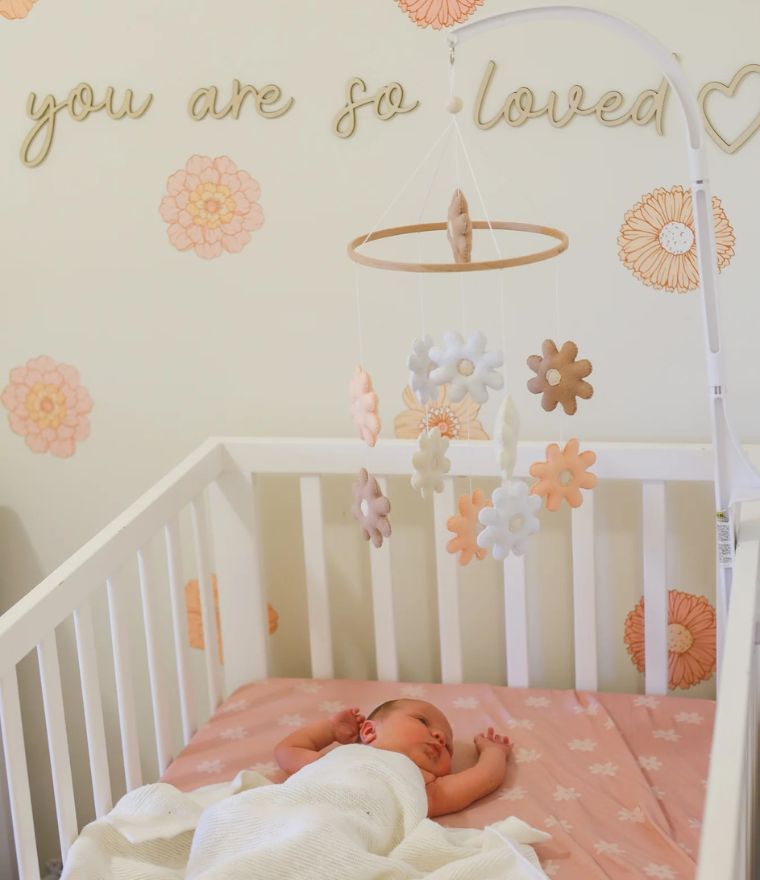 Daisy baby cot mobile with soft felt flowers above a sleeping newborn in a cozy nursery.