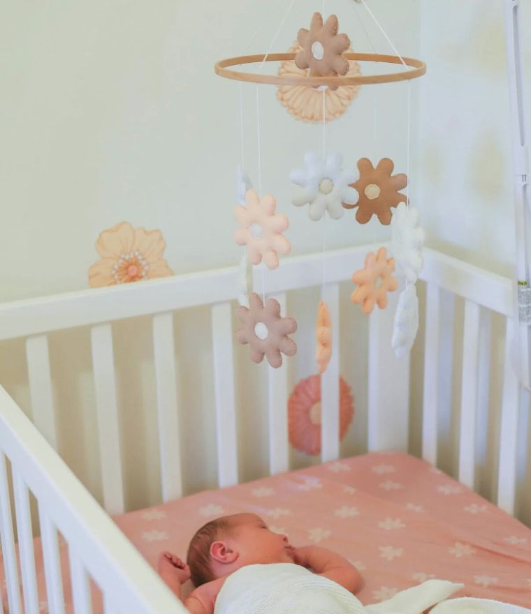 Daisy baby cot mobile in nursery above sleeping infant, featuring pastel felt daisies in neutral tones.