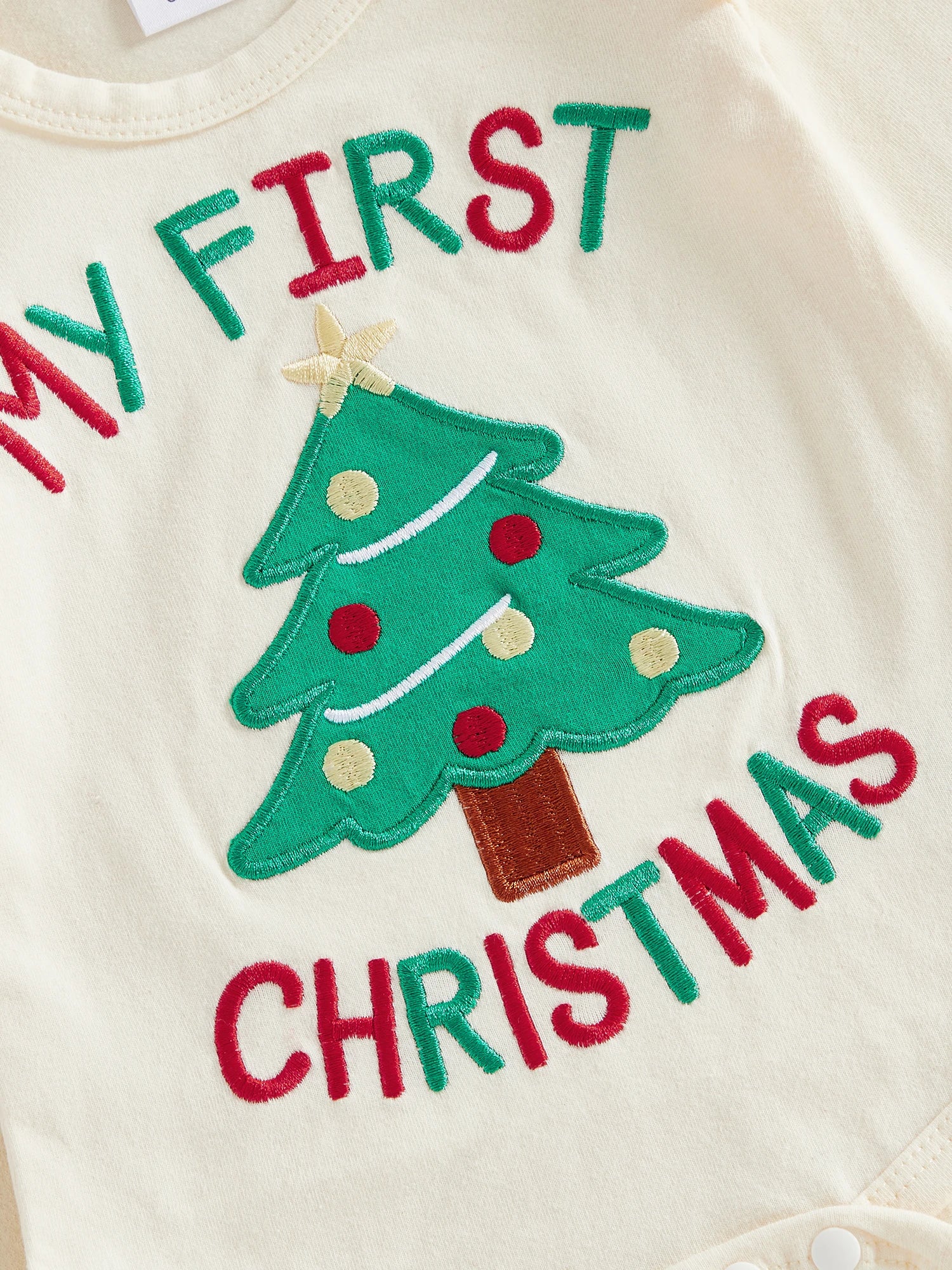 Close-up of ivory-coloured baby bodysuit featuring an embroidered 'MY FIRST CHRISTMAS' and a green Christmas tree design