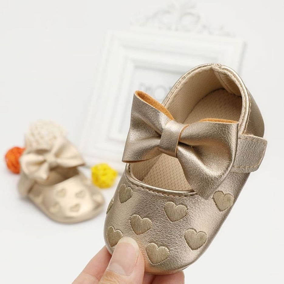 Gold baby moccasin with embossed hearts and bow held in hand showing size