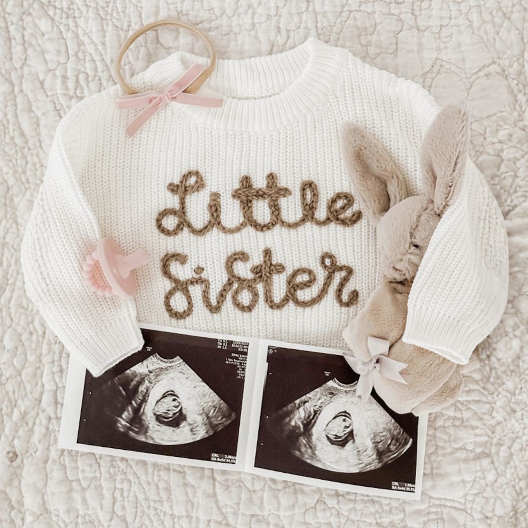 A white little sister jumper laid flat with ultrasound photos - baby pregnancy announcement