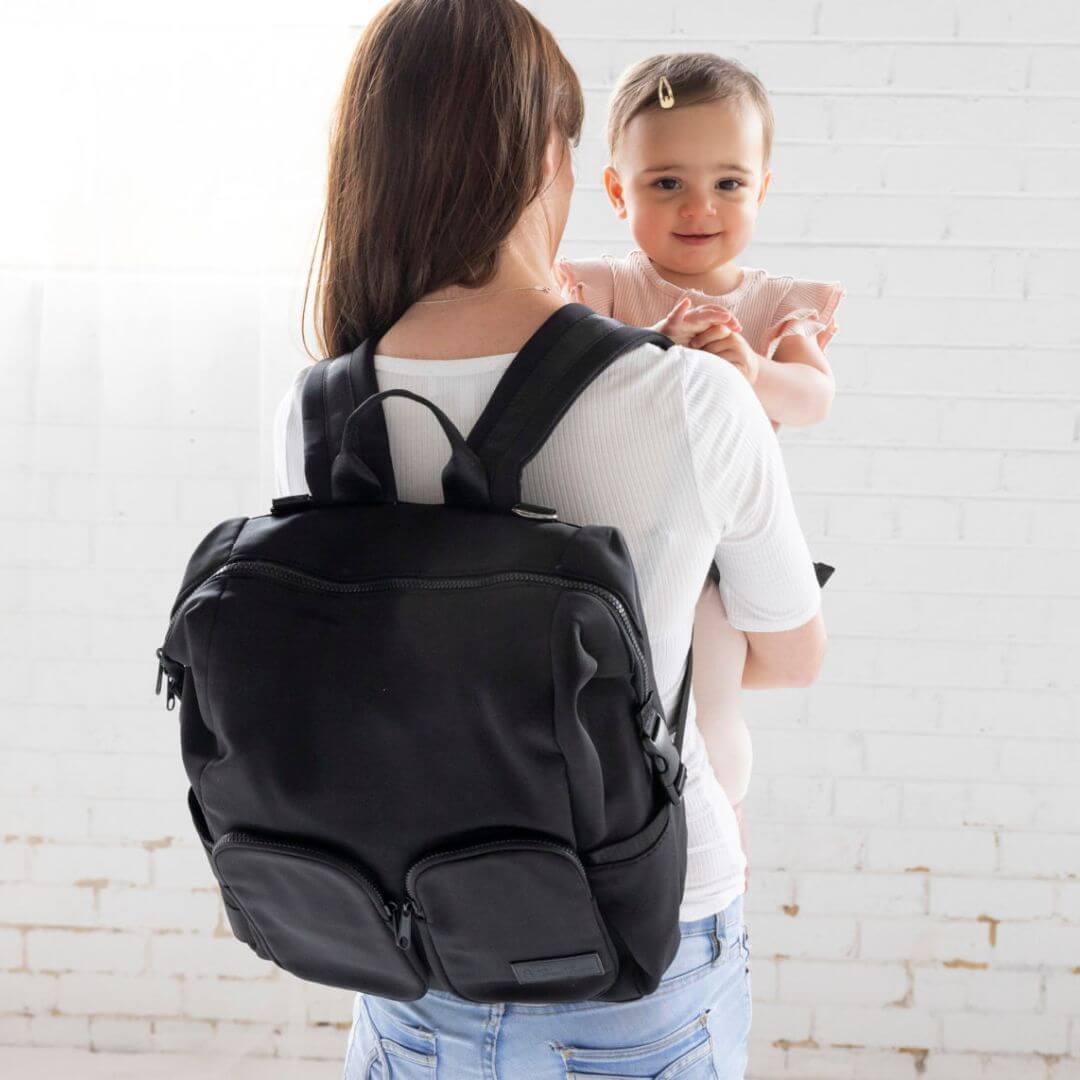 A mother wearing her black neoprene nappy backpack, holding a baby