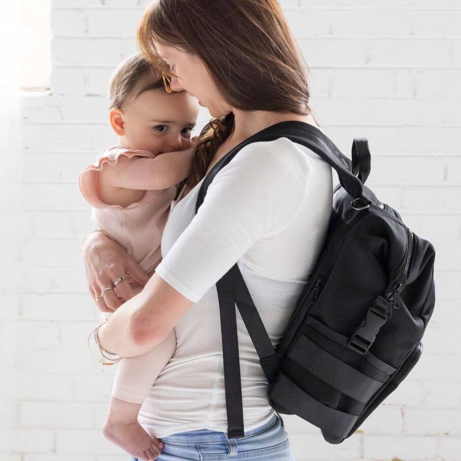 A mother wearing black neoprene nappy backpack, holding a baby - side view