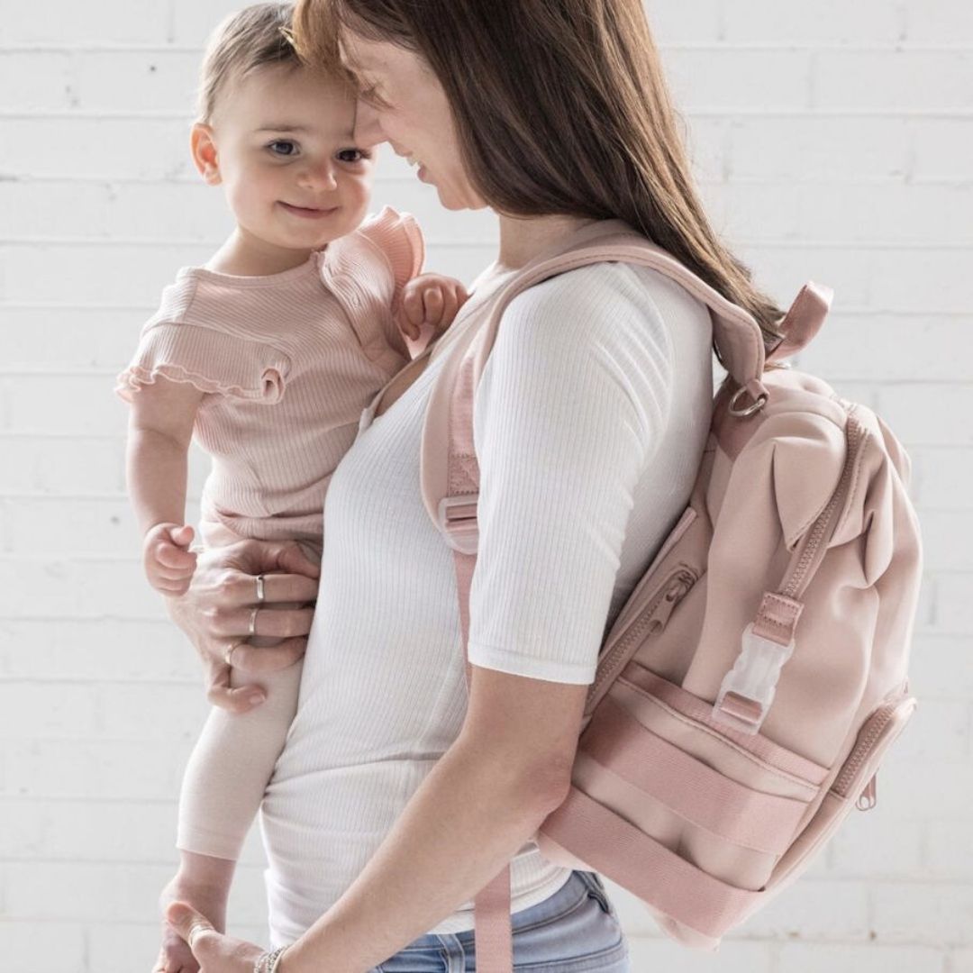 A mother wearing her blush pink neoprene nappy backpack, holding a baby - side view