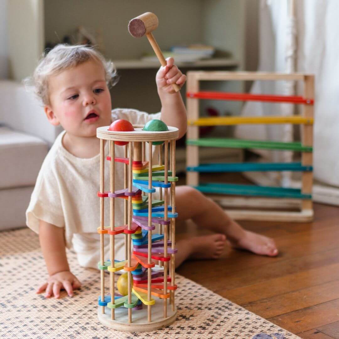 Toddler playing with wooden pound a ball tower with rainbow coloured curved tracks, three balls and wooden hammer