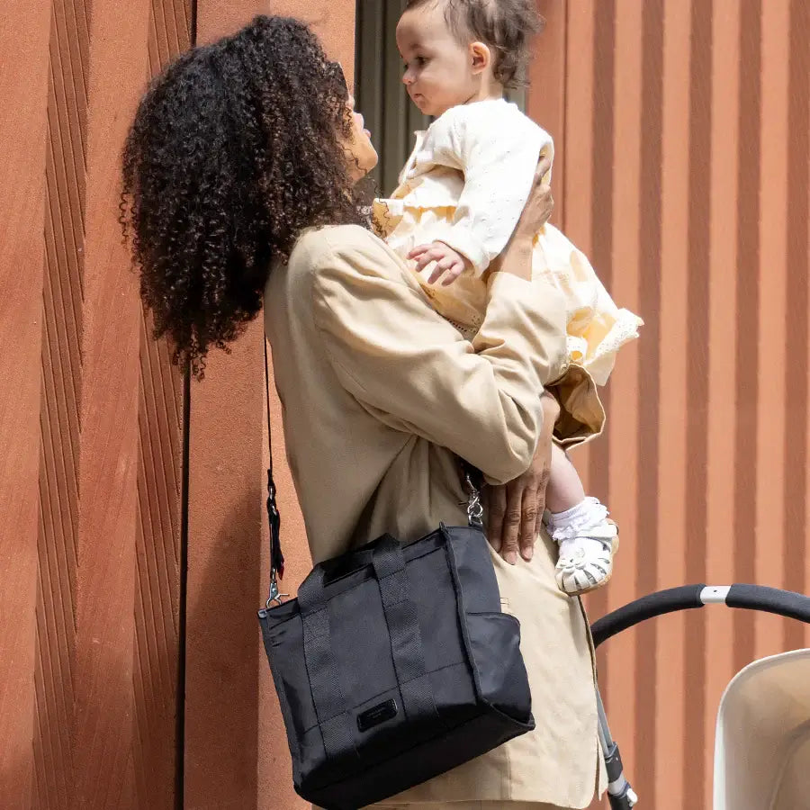 A mother holding her child while wearing the Storksak Thea Tote, showcasing practical parenting style.
