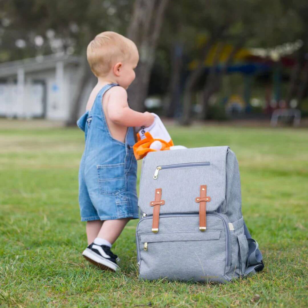 Baby standing next to grey nappy bag backpack in park