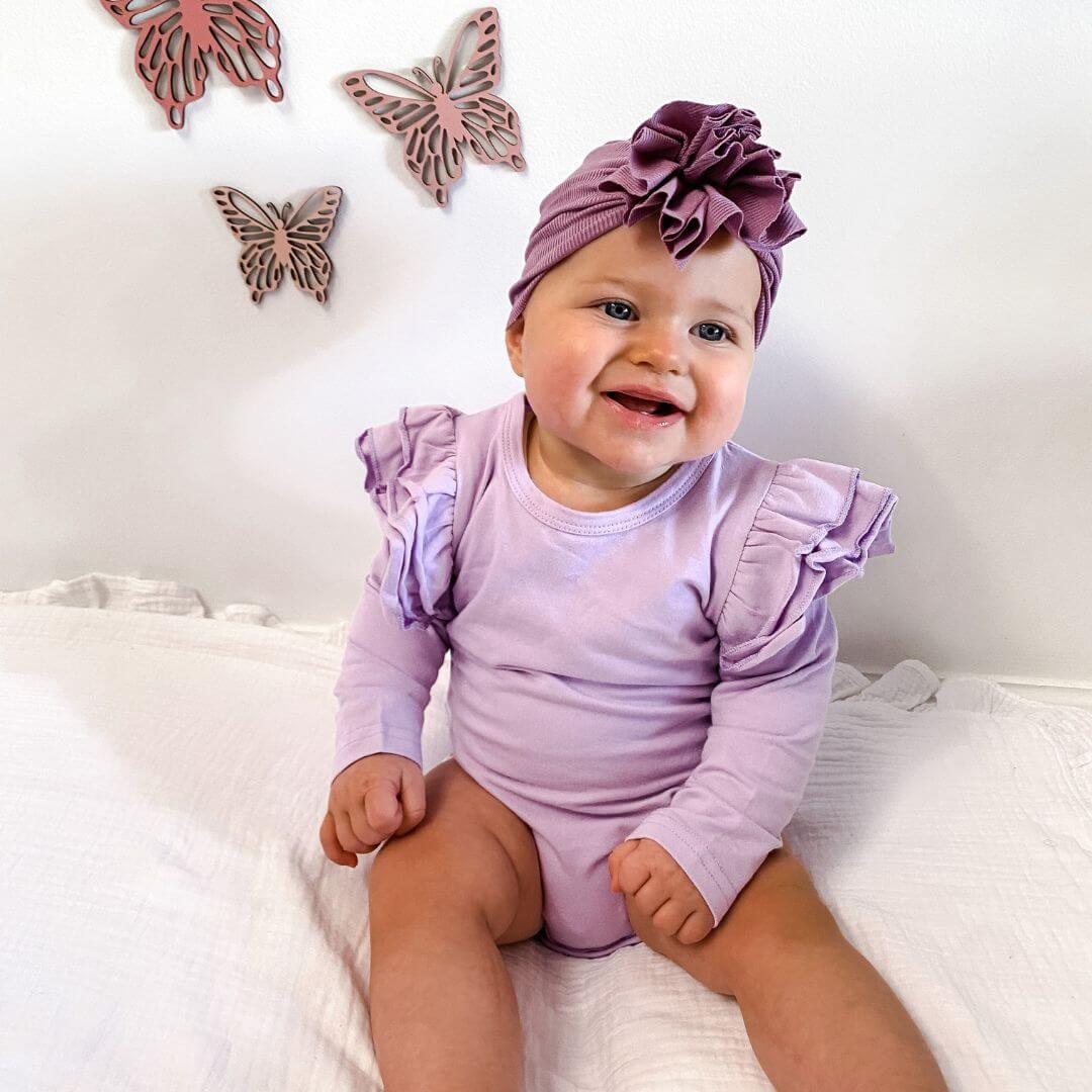 Smiling baby girl wearing lavender long sleeve flutter bodysuit and flower turban
